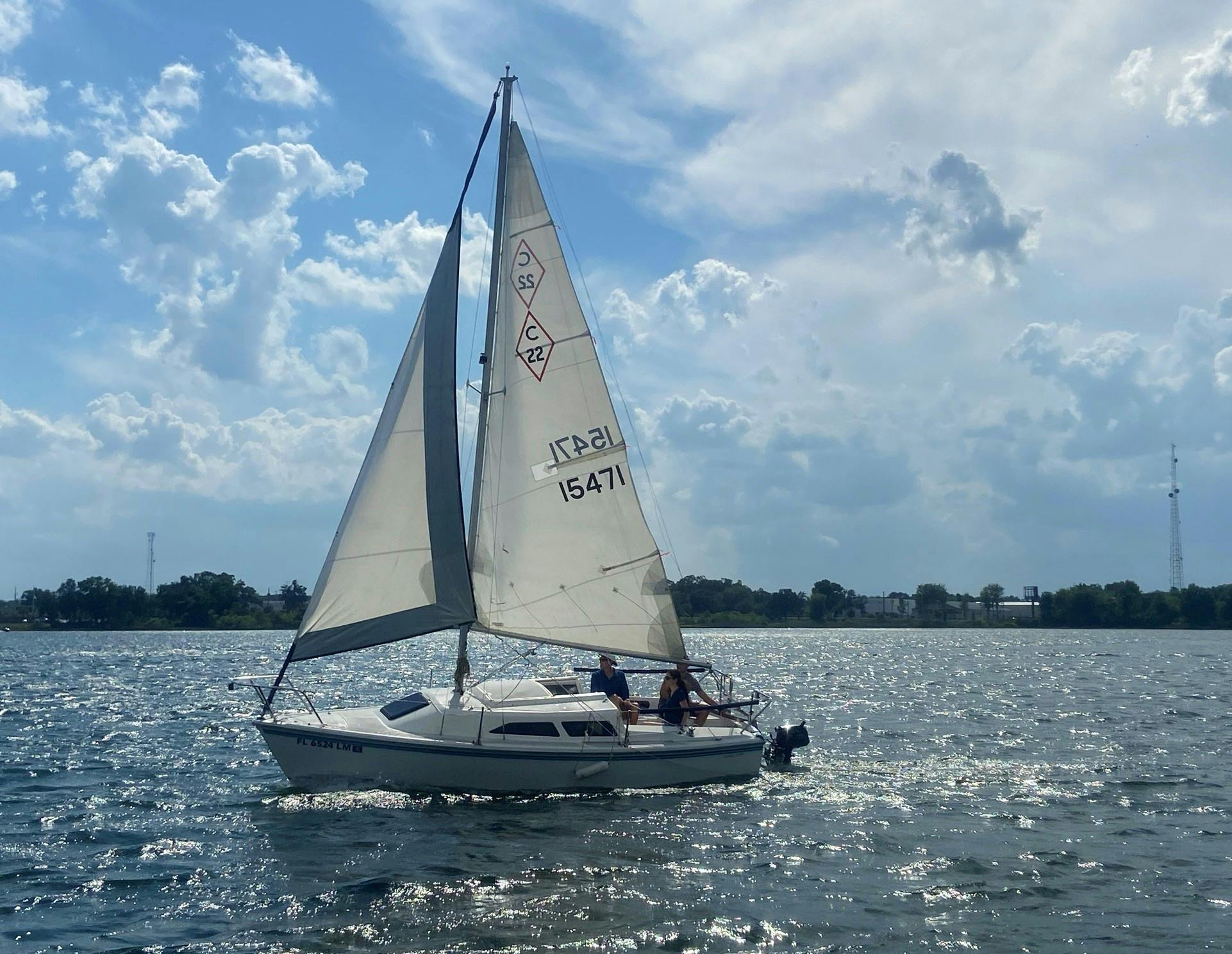 Un petit voilier aux voiles blanches sur un plan d'eau calme, sous un ciel partiellement nuageux, avec deux personnes à bord.