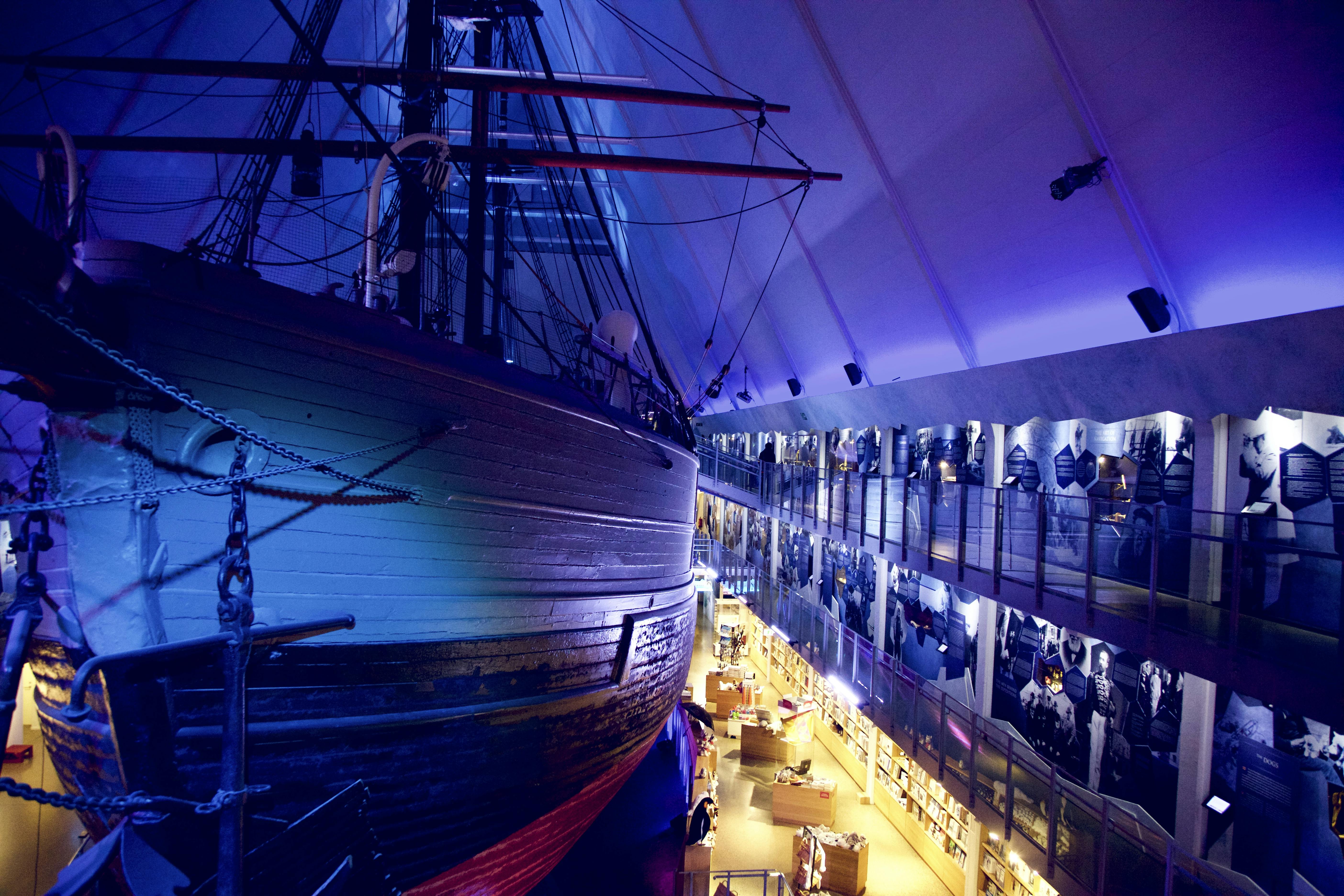 Μεγάλο ξύλινο πλοίο εκτίθεται σε εσωτερικούς χώρους κάτω από μπλε φωτισμό δίπλα σε μια γκαλερί δύο επιπέδων με ιστορικά εκθέματα και επισκέπτες.