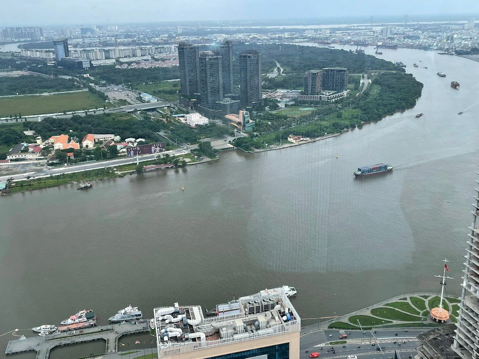 Aerial view of a river with boats, buildings, and green areas on the shore, and a cityscape in the distance.