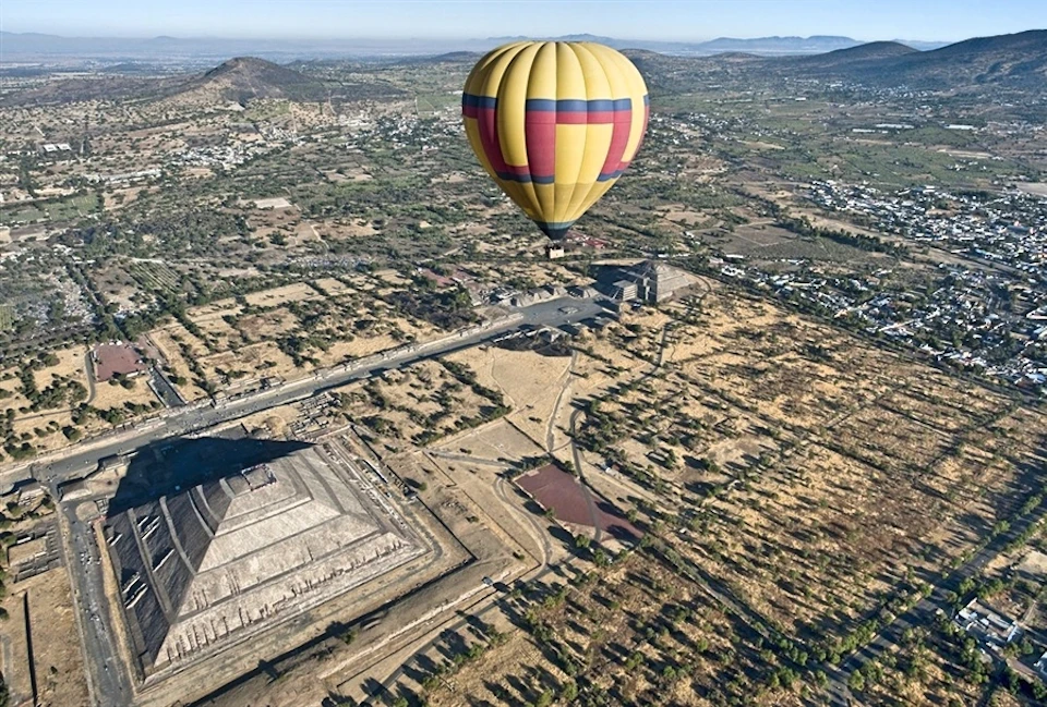 A hot air balloon floats above ancient pyramids and a sprawling landscape with sparse vegetation and distant mountains.
