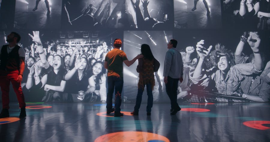 Tři lidé stojí před velkou obrazovkou s černobílým obrazem jásajícího koncertního davu.