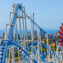 A blue and white rollercoaster with loops and drops, with a cityscape and ocean in the background under a clear sky.