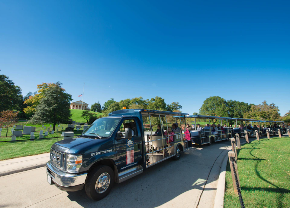 A tour tram full of passengers drives through a park with trees, grass, and a building in the background under a clear blue sky.