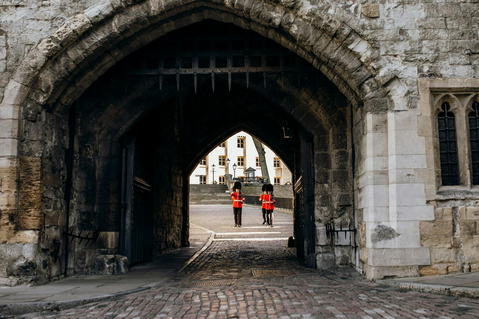 Two guards in red uniforms and black bearskin hats stand under a stone archway, with a historic building visible in the background.