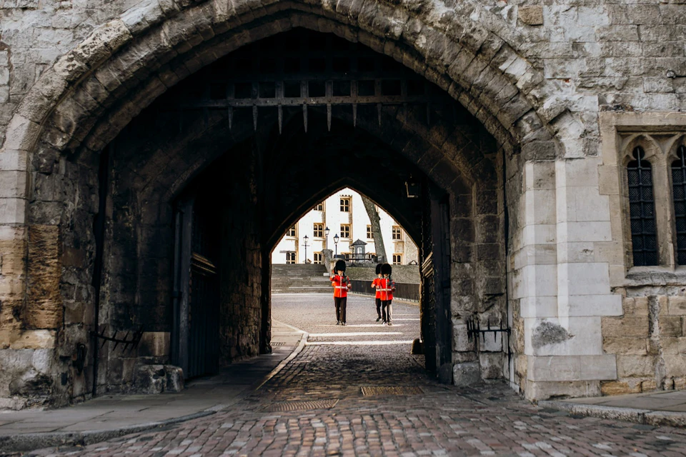 Two guards in red uniforms and black bearskin hats stand under a stone archway, with a historic building visible in the background.