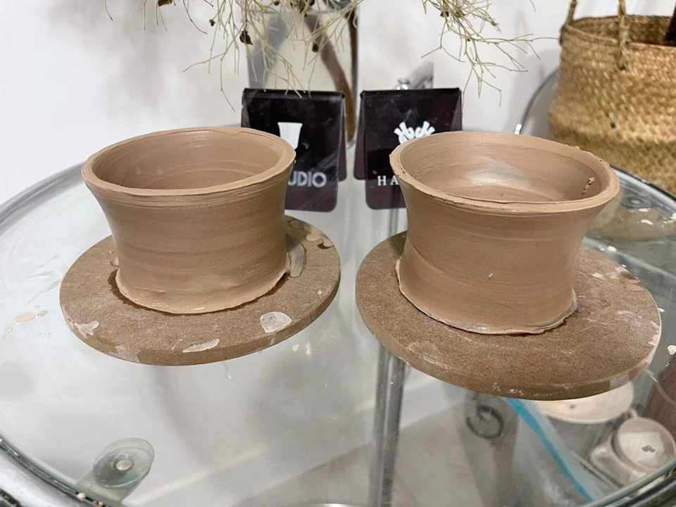 Two handmade clay pots on rotating platforms, partially finished, placed on a glass surface with tools in the background.