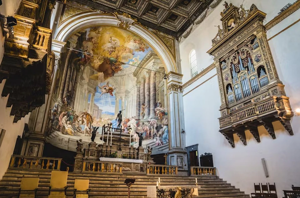 A grand church interior with a painted arched ceiling, ornate altar, intricate carvings, and a large pipe organ to the right.