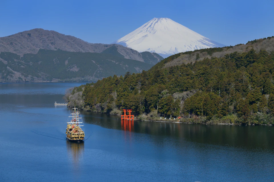 A scenic view of a lake with a traditional ship, a red torii gate on the shore, and a snow-capped mountain in the background.