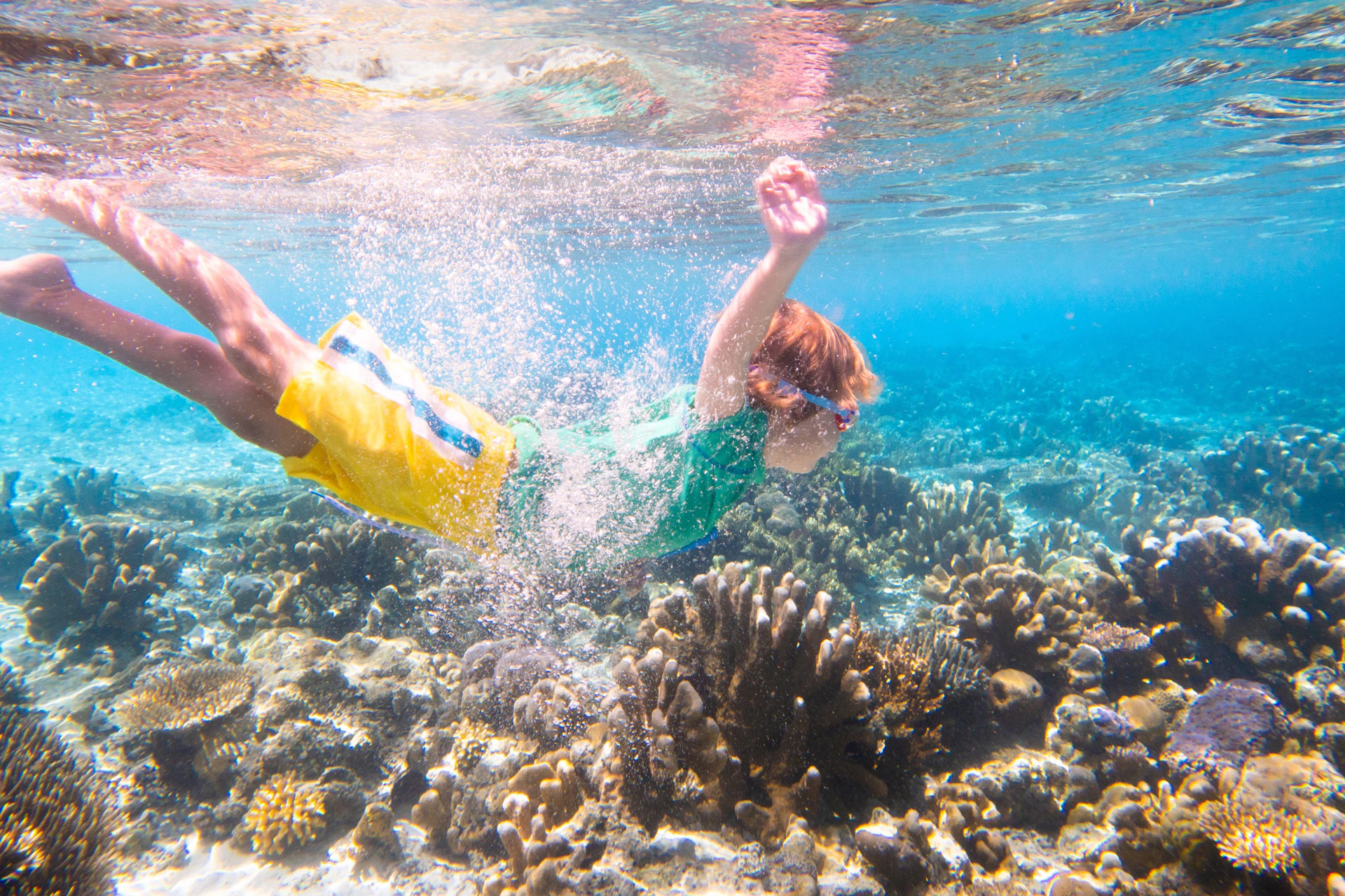 A person wearing yellow shorts and a green shirt swims underwater above coral in a clear and vibrant ocean scene.