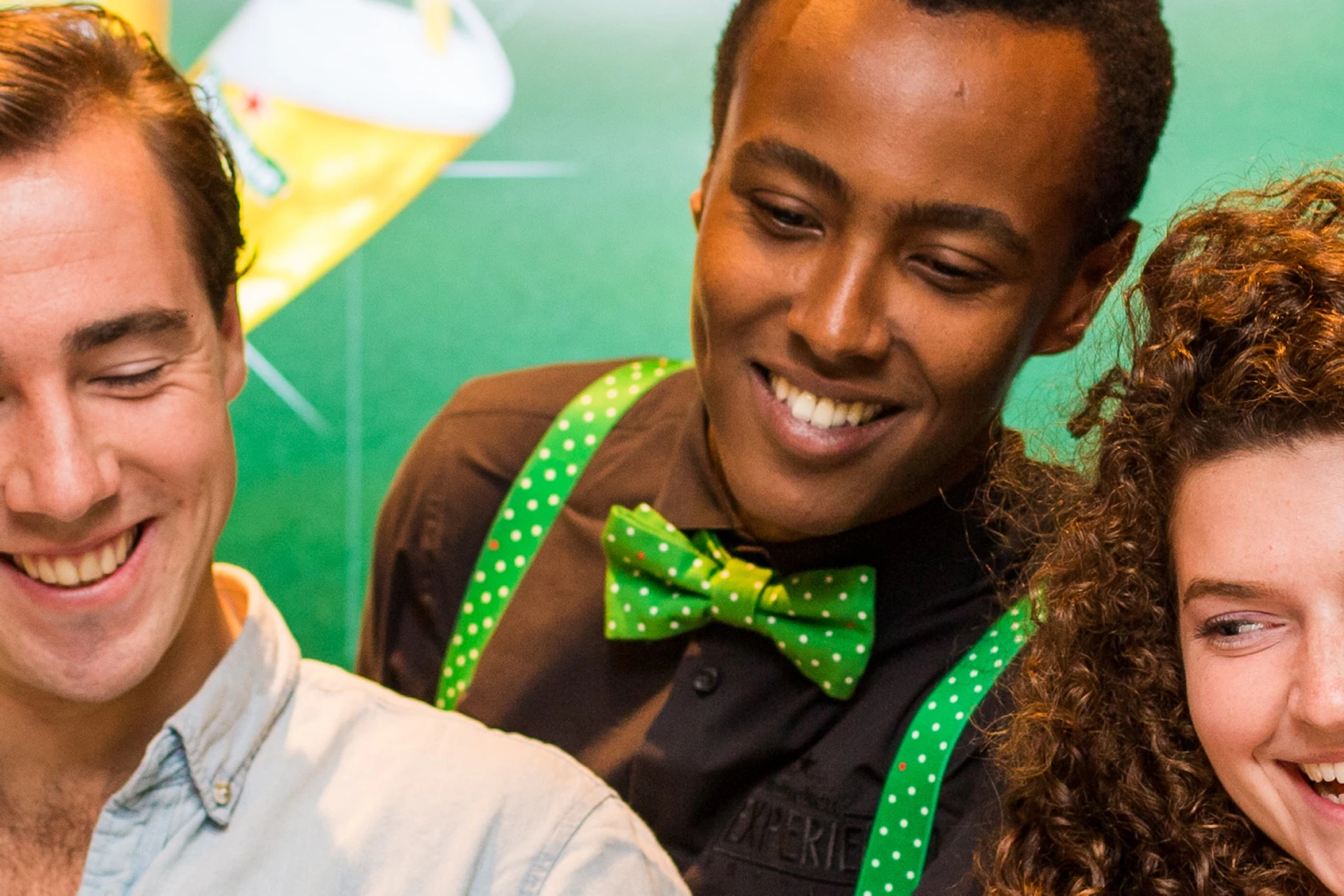 Three smiling people pouring beer from taps at a bar, with a green background and beer glasses in front of them.