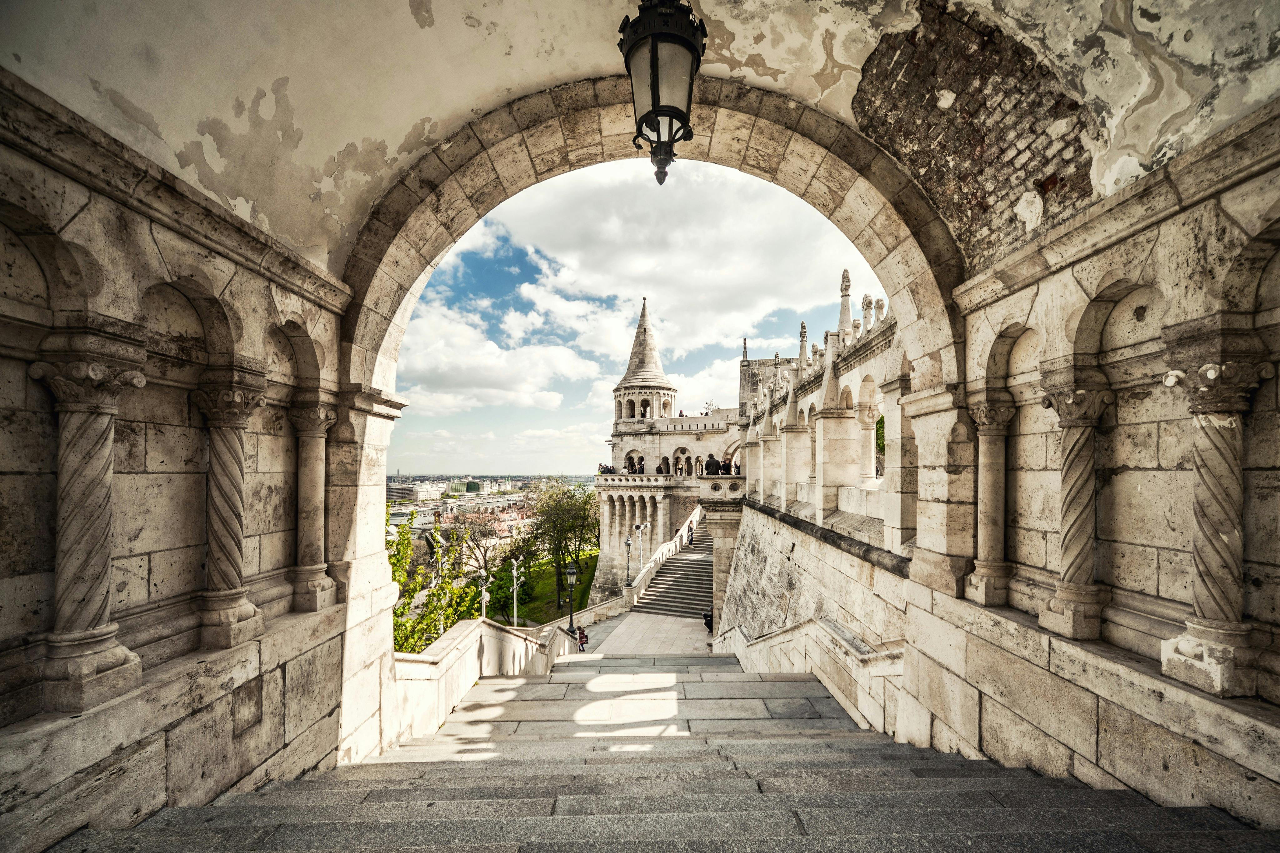 Stone archway framing a staircase leading to a historic European building with towers and a scenic view of the city.