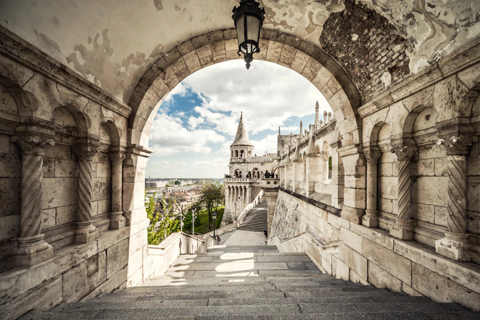 Stone archway framing a staircase leading to a historic European building with towers and a scenic view of the city.