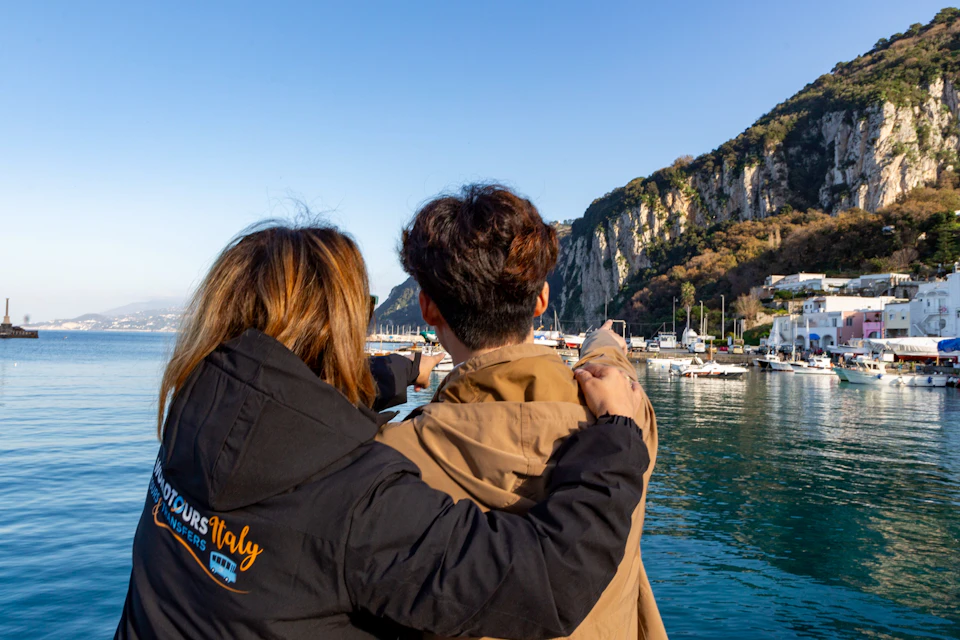 Two people stand by a waterfront, one pointing toward boats docked near a rocky cliff under a clear blue sky.