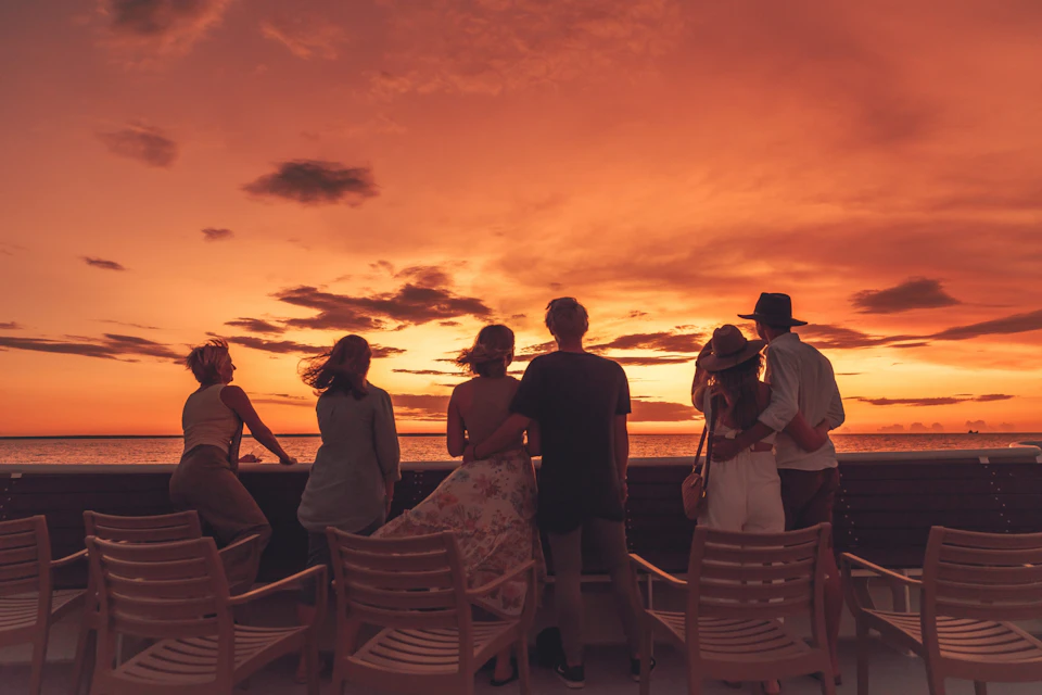 Six people stand and sit on a deck, watching a vibrant orange sunset over the ocean, with wooden chairs around them.