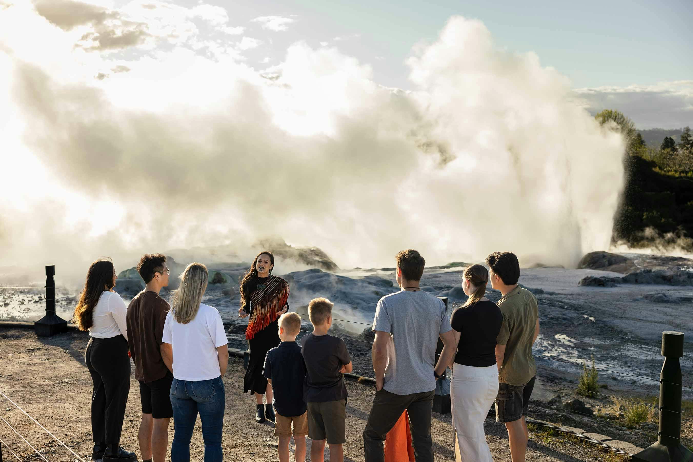 A group of people standing outdoors listening to a guide with steam rising from the ground in the background.