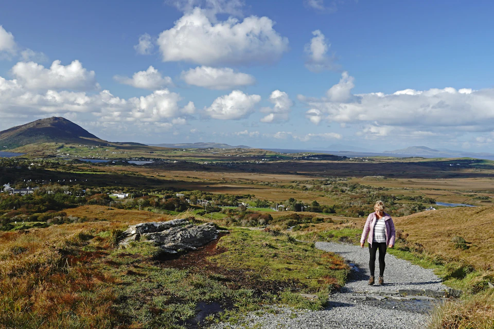 A woman walks along a gravel path in a vast, open landscape under a partly cloudy sky.