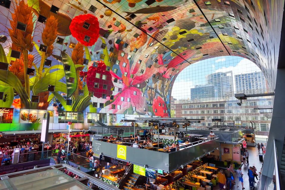 A vibrant, indoor market with colorful fruit and floral artwork on a curved ceiling. Shoppers and diners fill the multi-level space.