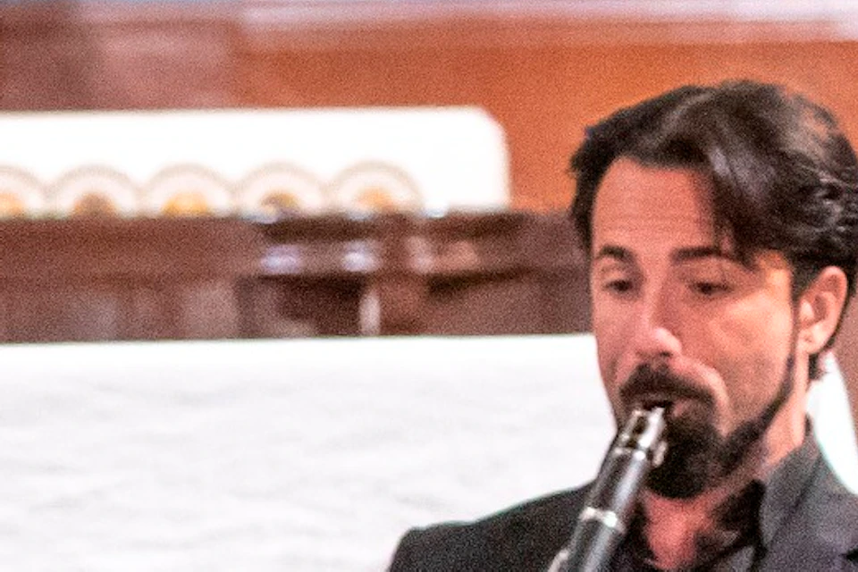 A man in a tuxedo sings on stage with musicians playing a flute, piano, and clarinet in an ornate venue.