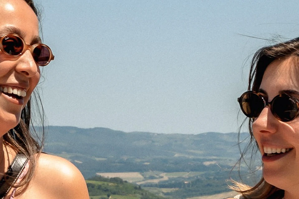 Two women sitting on a stone wall, smiling, with a scenic countryside background and a clear blue sky.