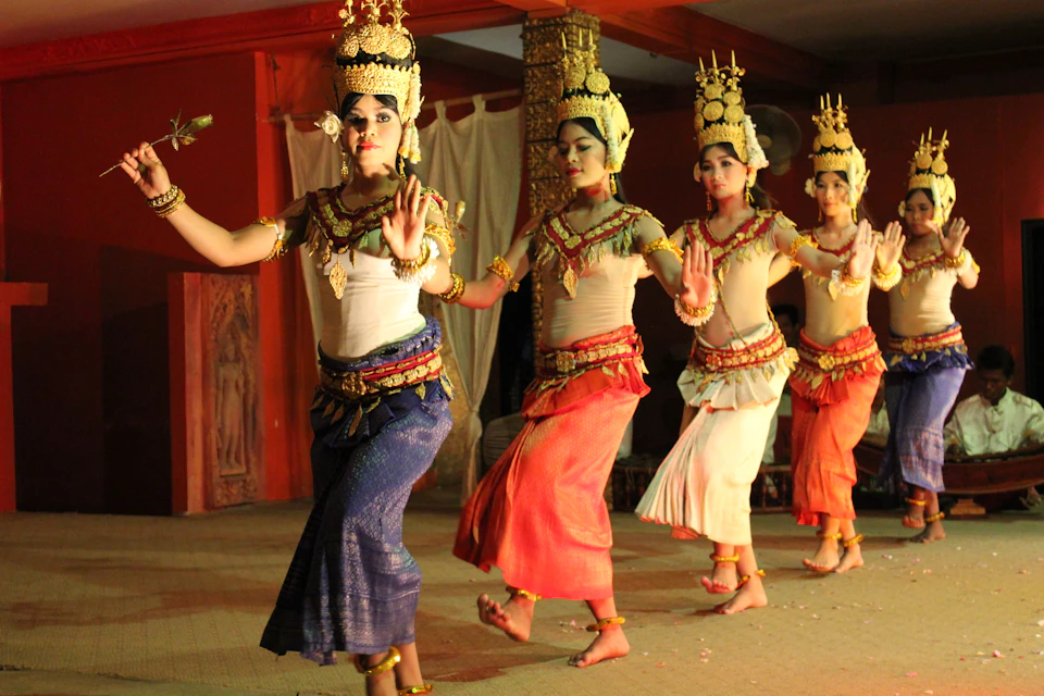 Four dancers in traditional attire with ornate headpieces perform in unison on a stage, with decorative pillars in the background.