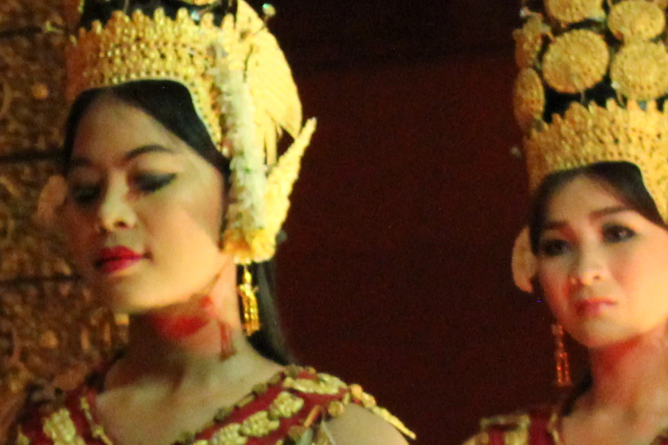 Four dancers in traditional attire with ornate headpieces perform in unison on a stage, with decorative pillars in the background.