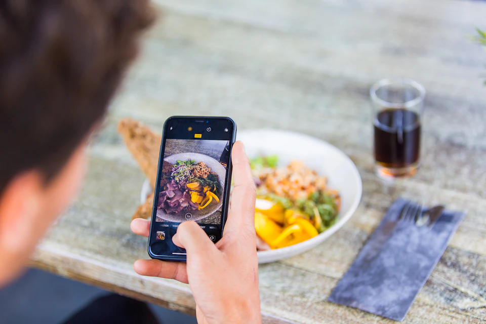 Person holding a smartphone, taking a photo of a plated meal on a wooden table. A glass with a dark beverage is nearby.