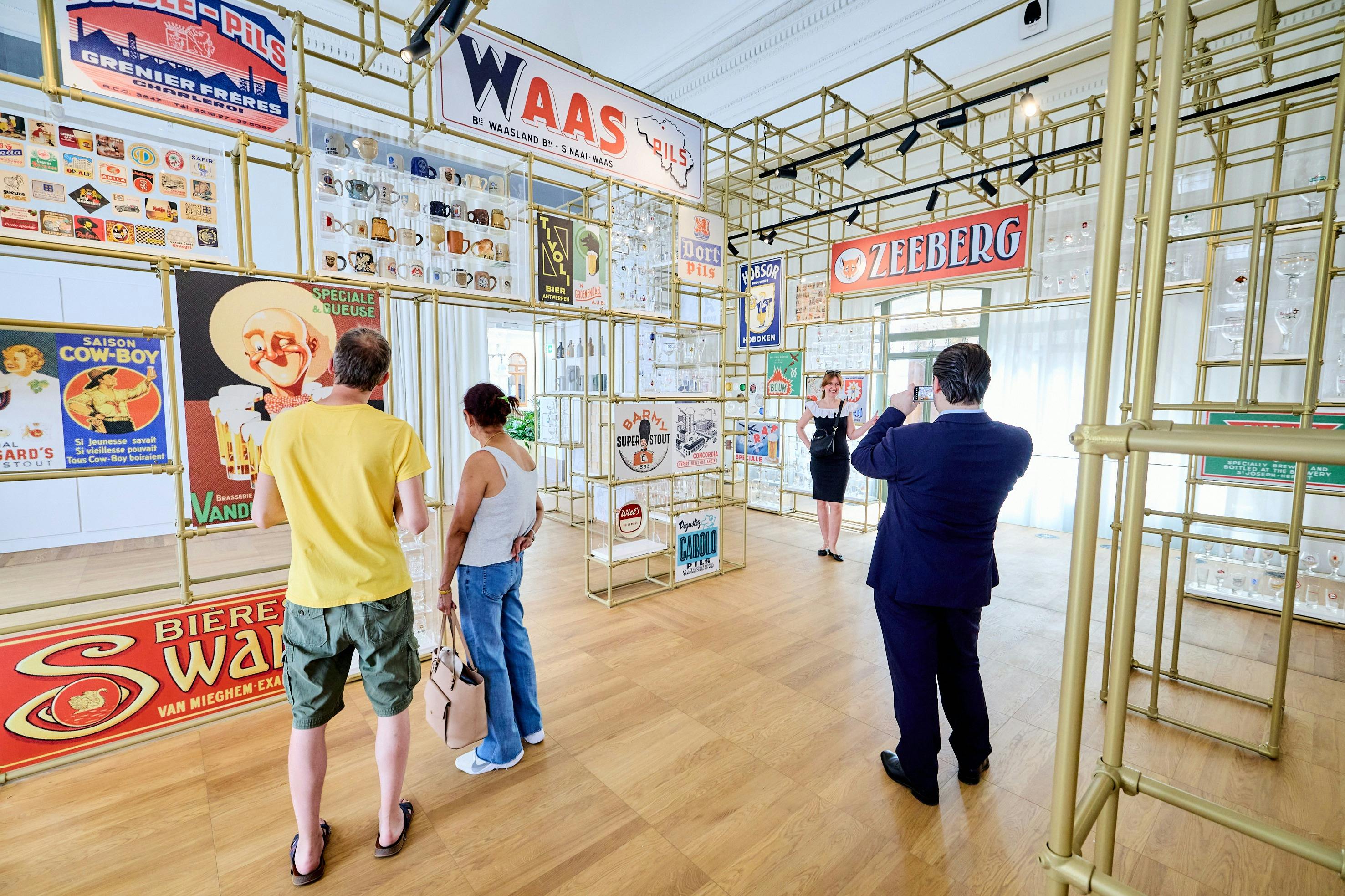 Lidé si prohlížejí vintage pivní cedule a upomínkové předměty v jasně osvětlené výstavní místnosti s dřevěnými podlahami a zlatou konstrukcí.