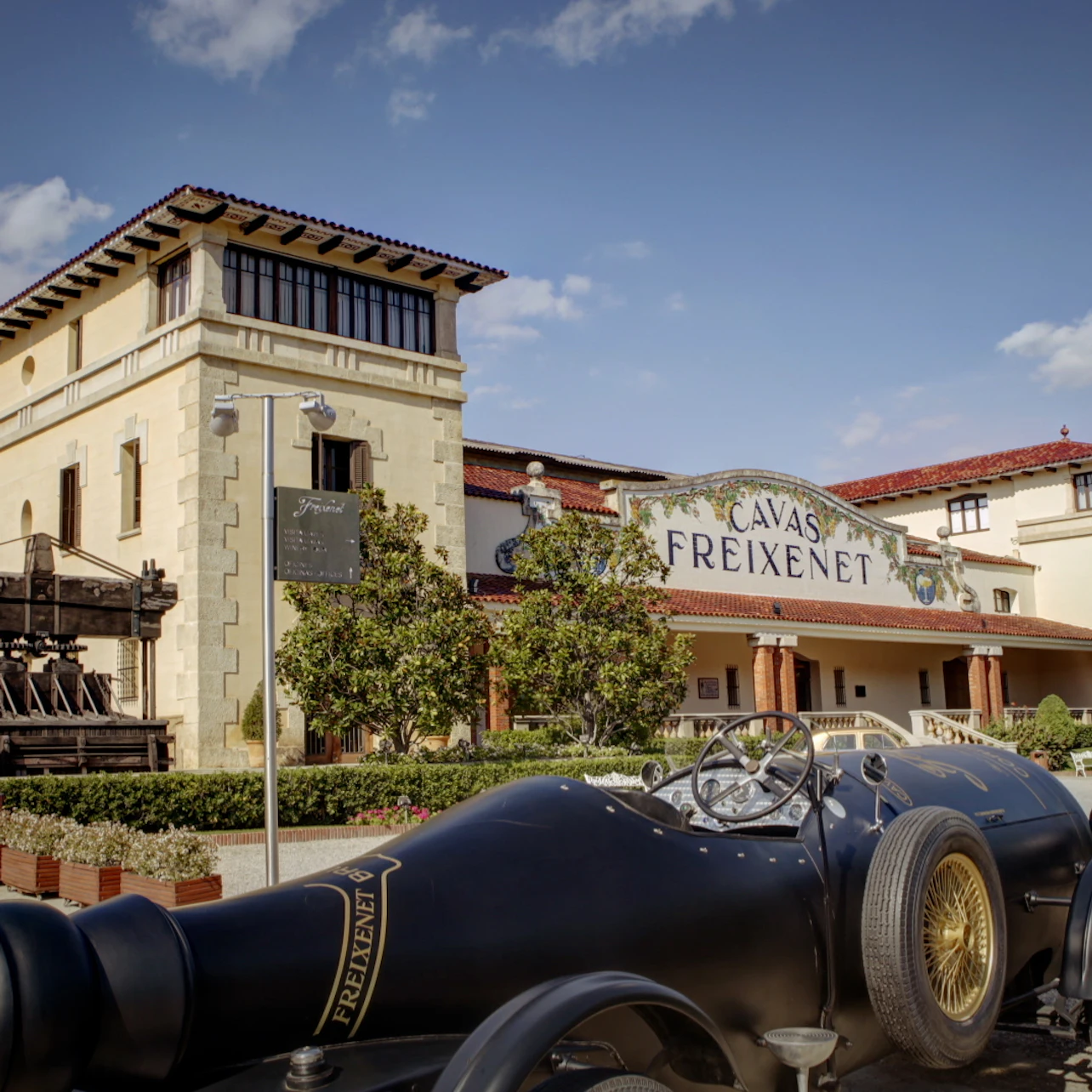 Utsidan av Cavas Freixenet-byggnaden med en veteranbil parkerad framför, grönska och klarblå himmel.