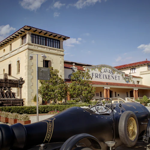 Utsidan av Cavas Freixenet-byggnaden med en veteranbil parkerad framför, grönska och klarblå himmel.