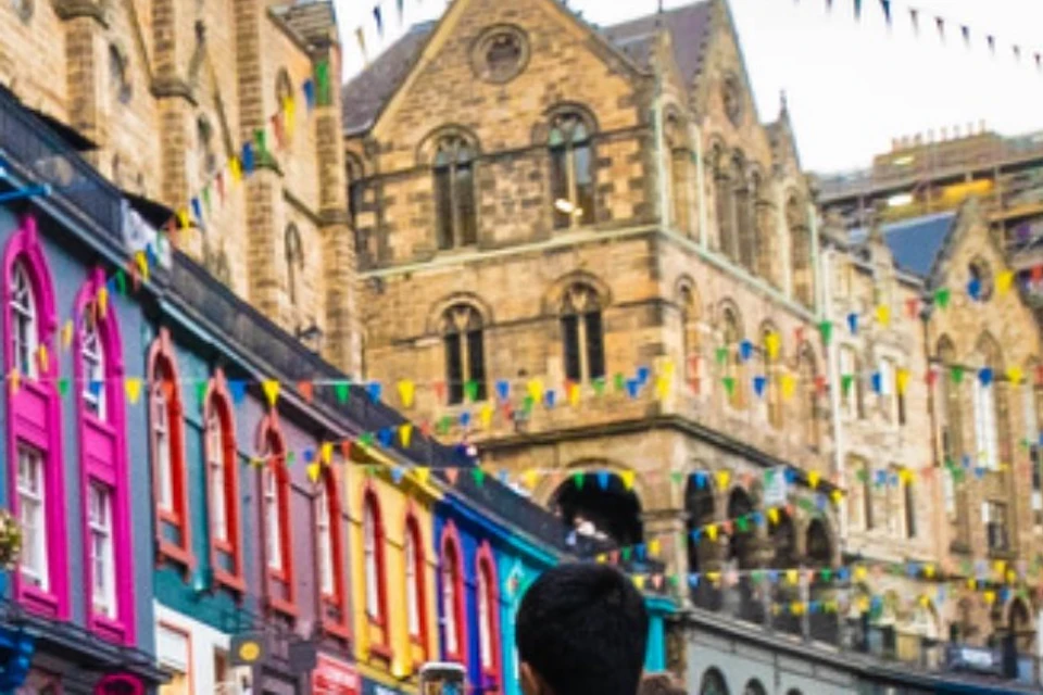 A woman in a brown coat smiles on a colorful cobblestone street lined with shops and buildings, with people and flags around.