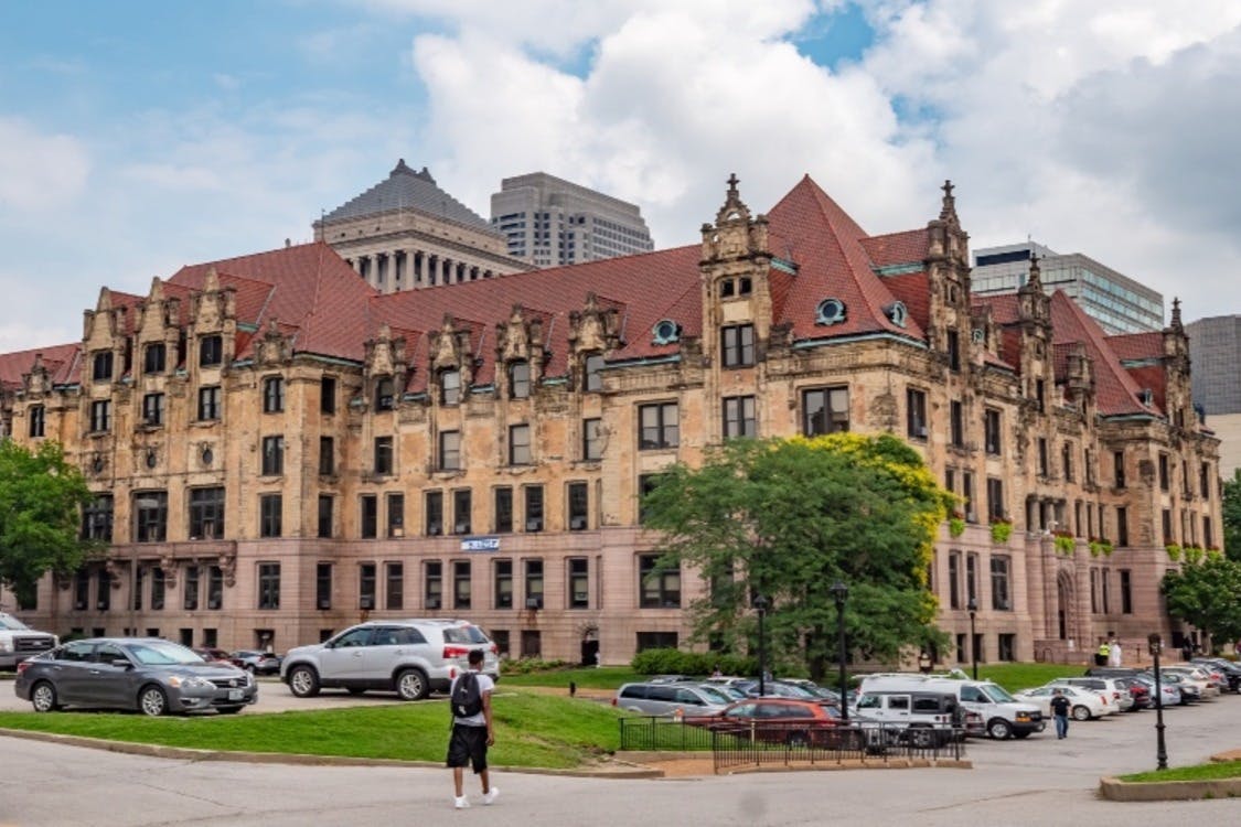 Ιστορικό κτίριο με κόκκινες κεραμοσκεπές, περιτριγυρισμένο από παρκαρισμένα αυτοκίνητα και λίγα δέντρα, με φόντο σύγχρονους ουρανοξύστες.