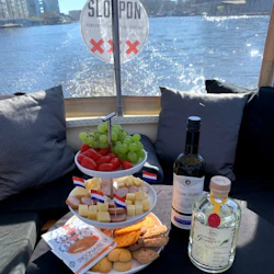 Snacks, vin och sprit på ett bord inuti en båt med utsikt över en vattenväg och "Sloep On Amsterdam Canal Tours" -logotypen.