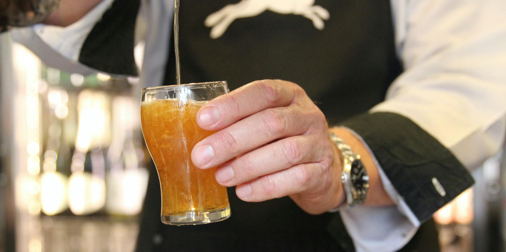 A hand holding a small glass of amber-colored liquid being poured, with a watch visible on the wrist of the hand.
