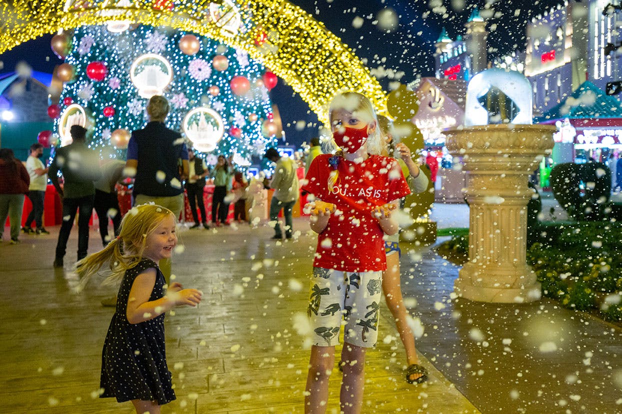 어린이들은 크리스마스 트리를 배경으로 축제 빛 장식 아래에서 인조 눈을 맞으며 놀고 있습니다.