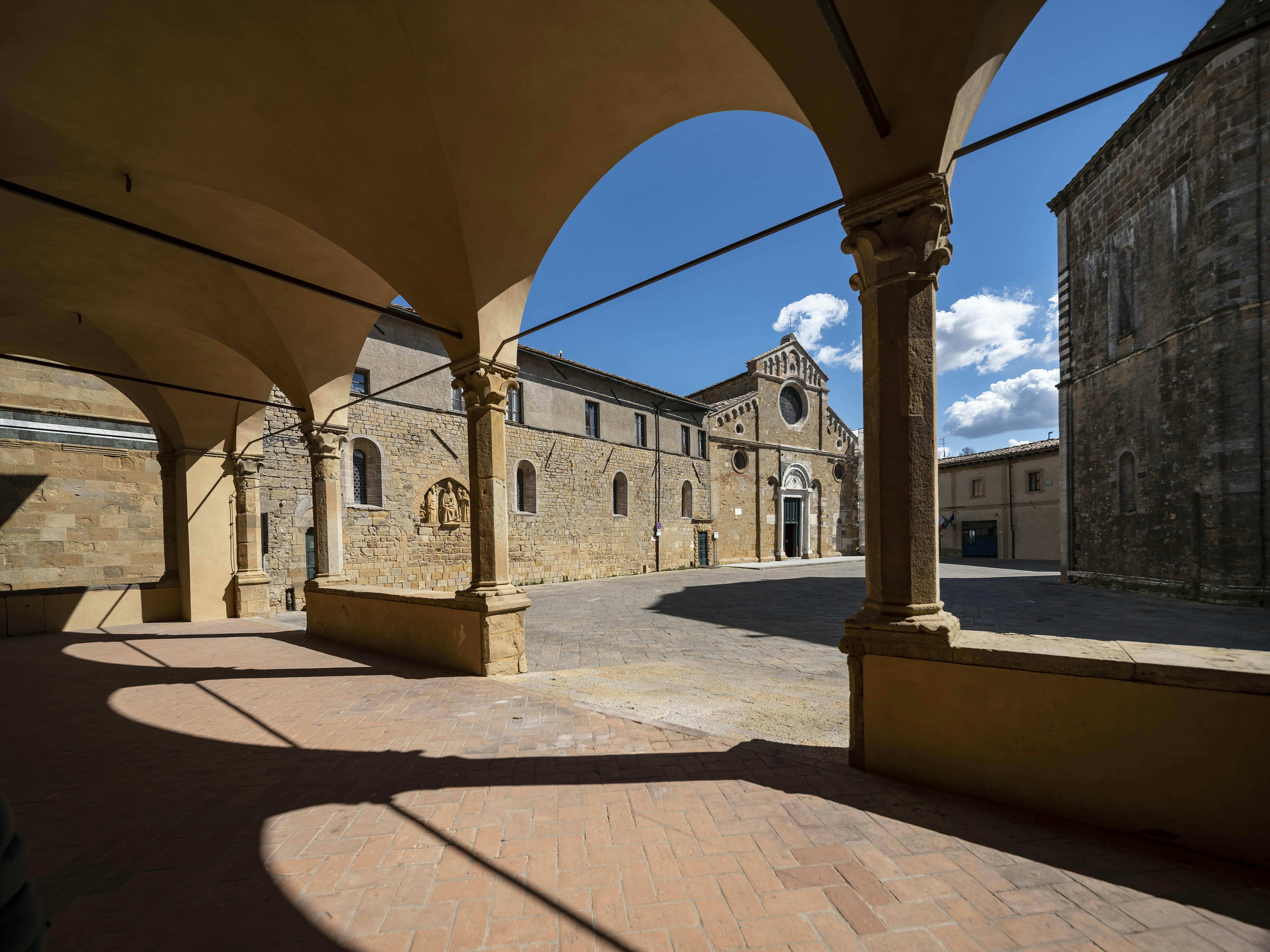 Un cortile in pietra con archi e un edificio storico con un rosone sotto un cielo blu con nuvole.