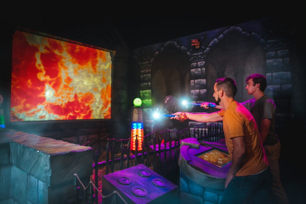 Two people in a dimly lit room aim glowing wands at targets on a game setup with stone walls and vivid lights.