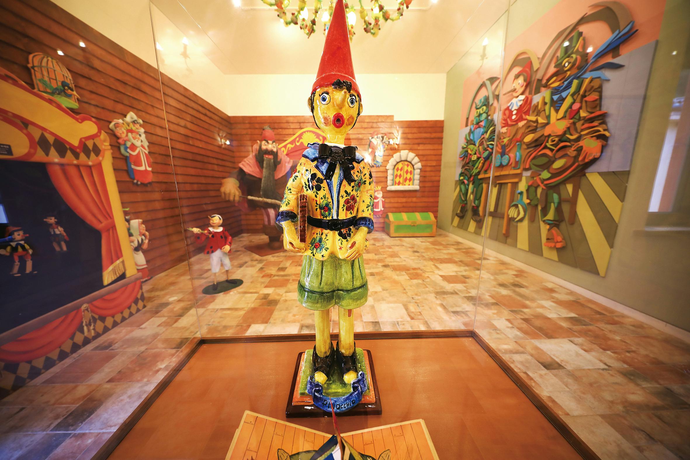 A colorful Pinocchio statue in a glass case, surrounded by a room with wooden walls and vibrant murals depicting scenes.