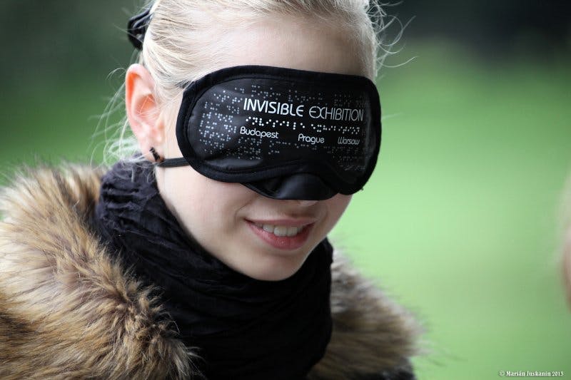 Une personne portant un bandeau noir étiqueté "EXPOSITION INVISIBLE" avec les villes de Budapest, Prague et Varsovie écrites dessus.