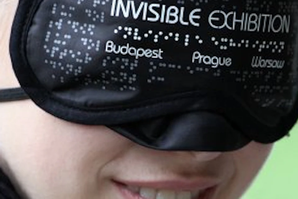 A person wearing a black blindfold labeled "INVISIBLE EXHIBITION" with cities Budapest, Prague, and Warsaw written on it.