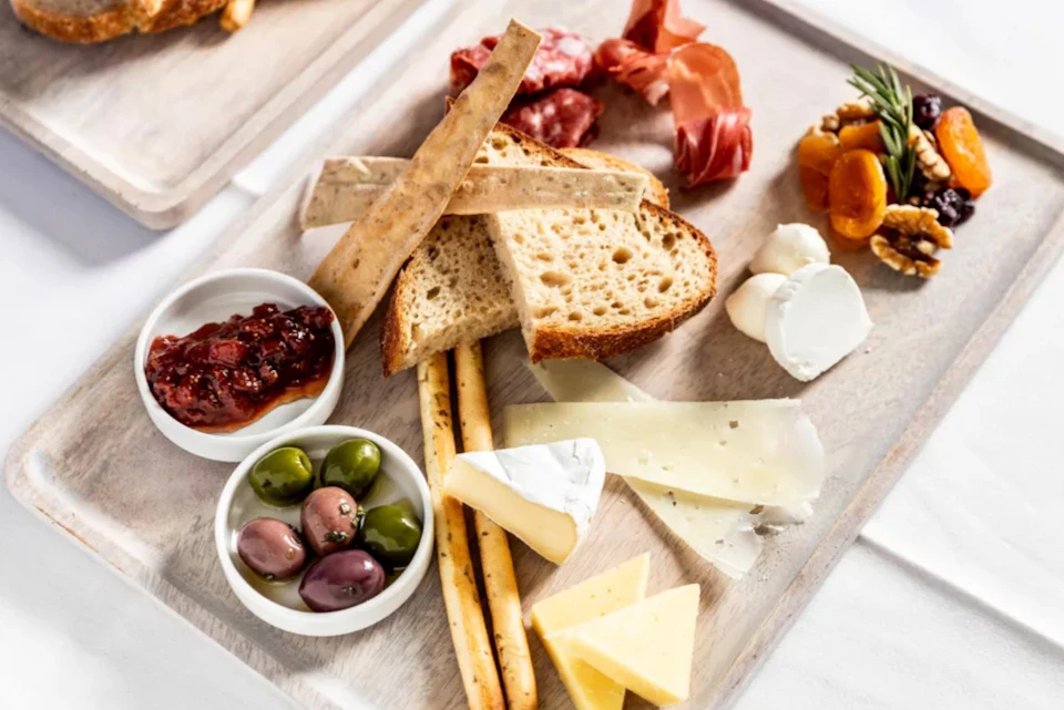 A charcuterie board featuring bread slices, cheeses, cured meats, olives, jam, and dried fruits on a light wooden surface.