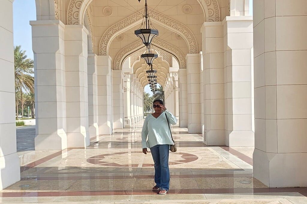 Woman poses in a corridor with ornate arches and hanging lanterns, standing on a polished floor with geometric designs.