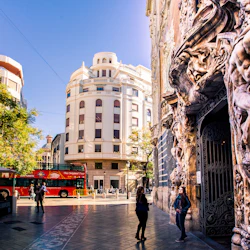 Gatuscen med utsmyckade byggnadsristningar, röd turnébuss och människor som går. Bleka byggnader och grönska i bakgrunden.