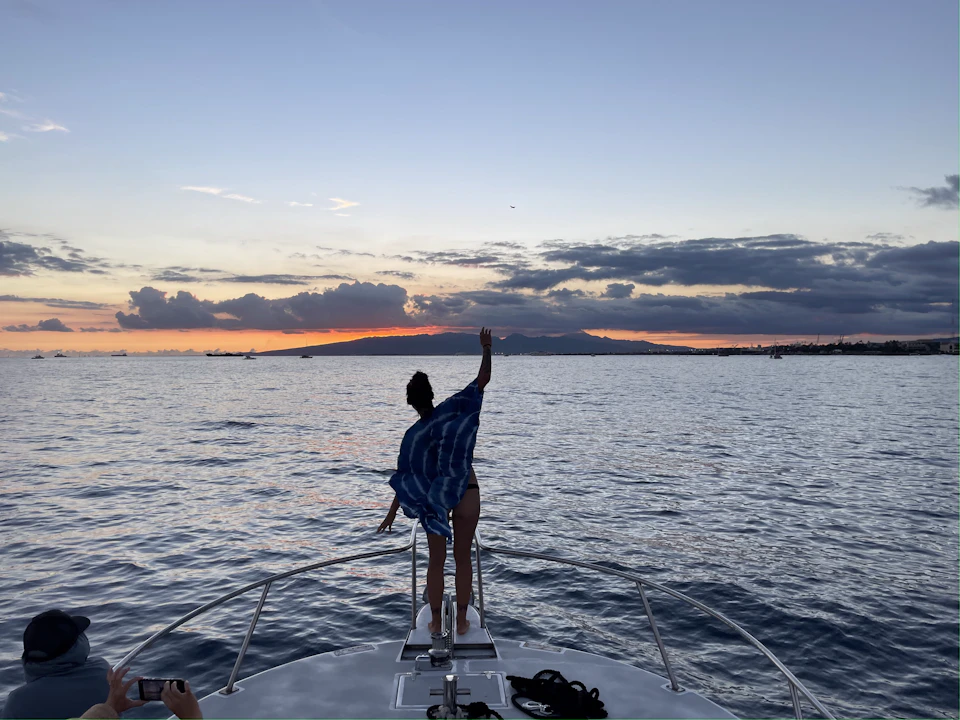 A person stands at the bow of a boat, facing a sunset over water, with their arm raised. Mountains and clouds are visible in the distance.