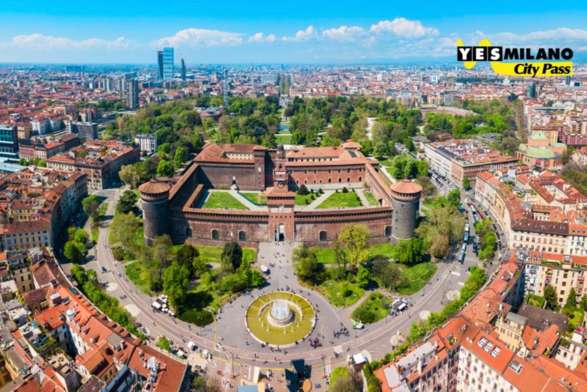 Luftaufnahme eines großen, historischen Backsteinschlosses mit vier Ecktürmen, umgeben von einer Ringstraße und einem grünen Park, in einer Stadt.