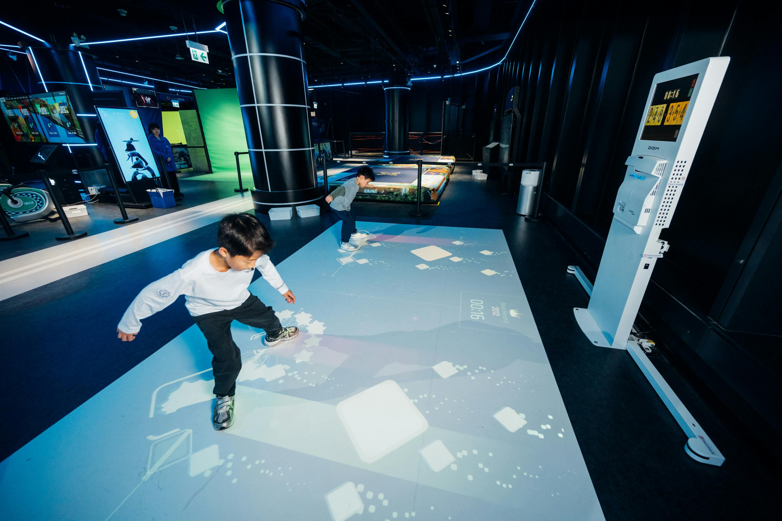 Two children interacting with a projected interactive floor display in a modern, dark-themed exhibition space.