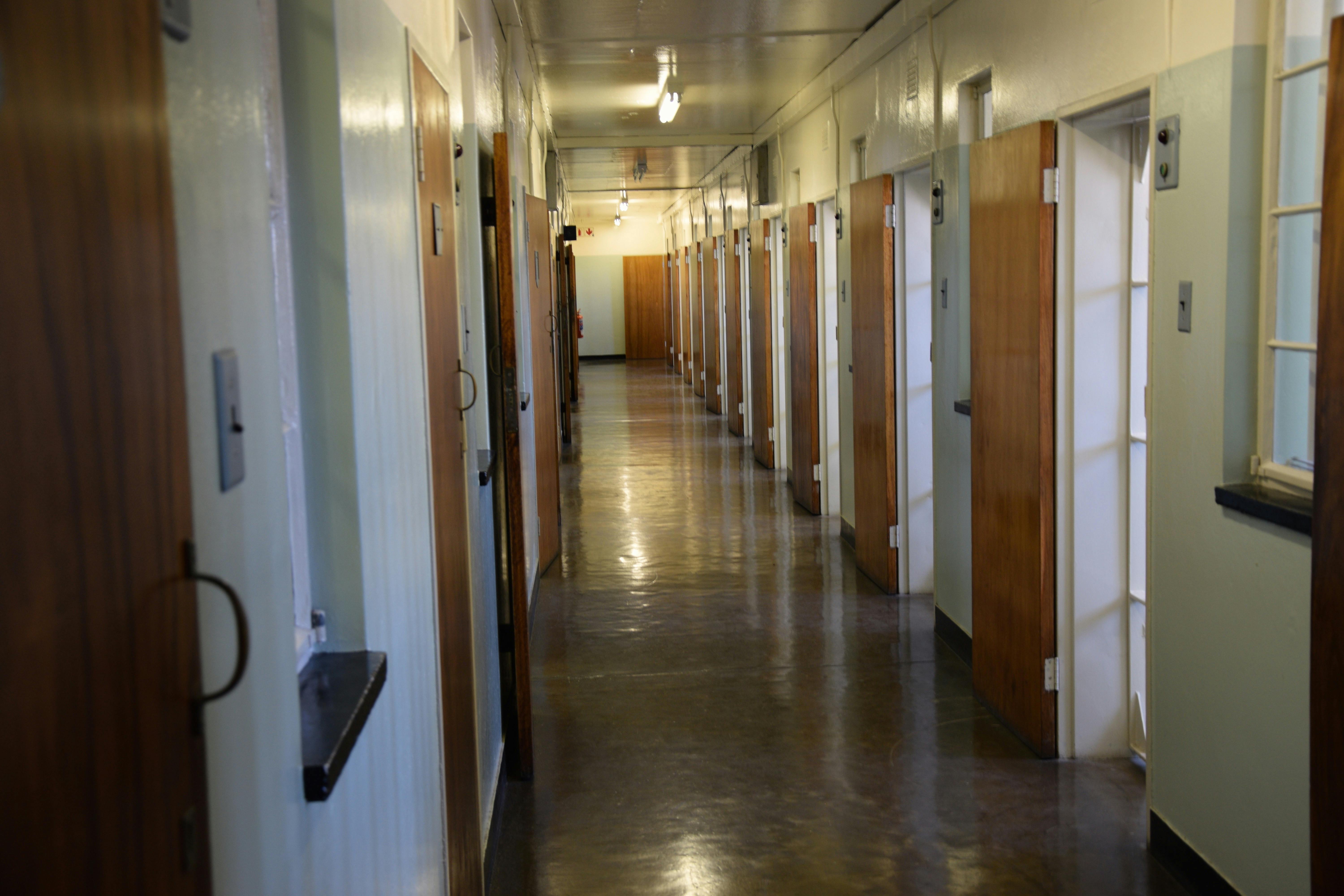 Dlouhá, prázdná chodba s několika dřevěnými dveřmi mírně pootevřenými a zářivkovým osvětlením nad hlavou.