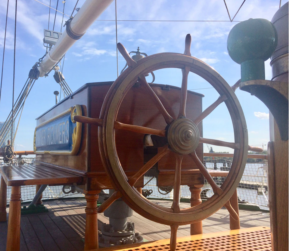 Wooden ship's wheel on the deck of a sailboat with ropes and rigging in the background under a partly cloudy sky.