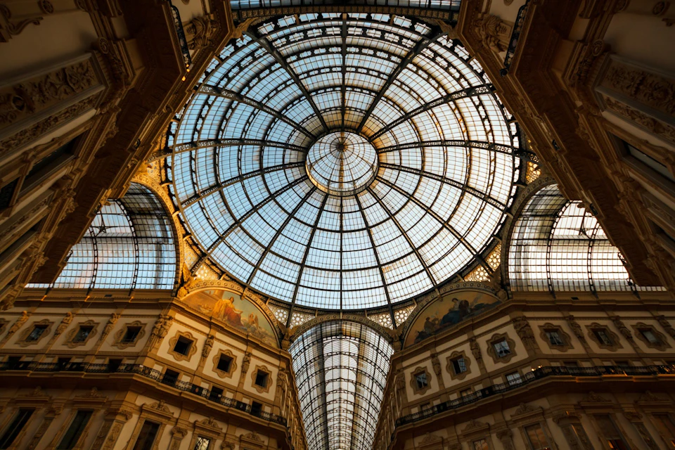 A glass dome ceiling with intricate framework, surrounded by ornate building facades in a historic architectural setting.