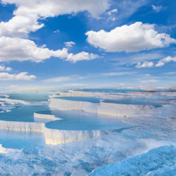 Terrasserade vita mineralpooler med klart blått vatten under en klarblå himmel med spridda moln.