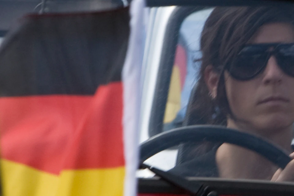 Red and silver vintage cars with German flags, driven by a woman and a man respectively, in a roadside procession.